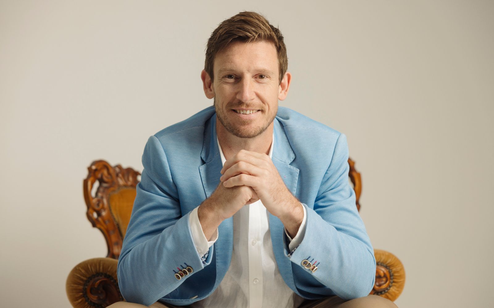 Mat Bankes seated in a carved timber chair, leaning forward with his hands clasped, smiling